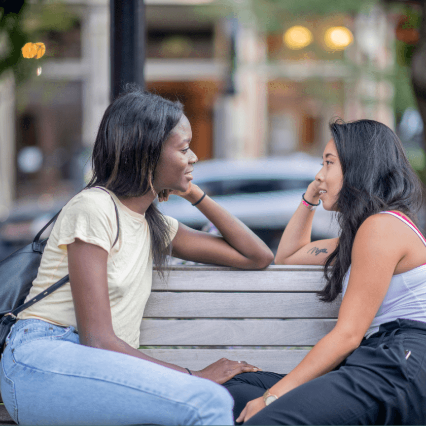 A lesbian interracial couple look lovingly into each other's eyes while sitting on a bench